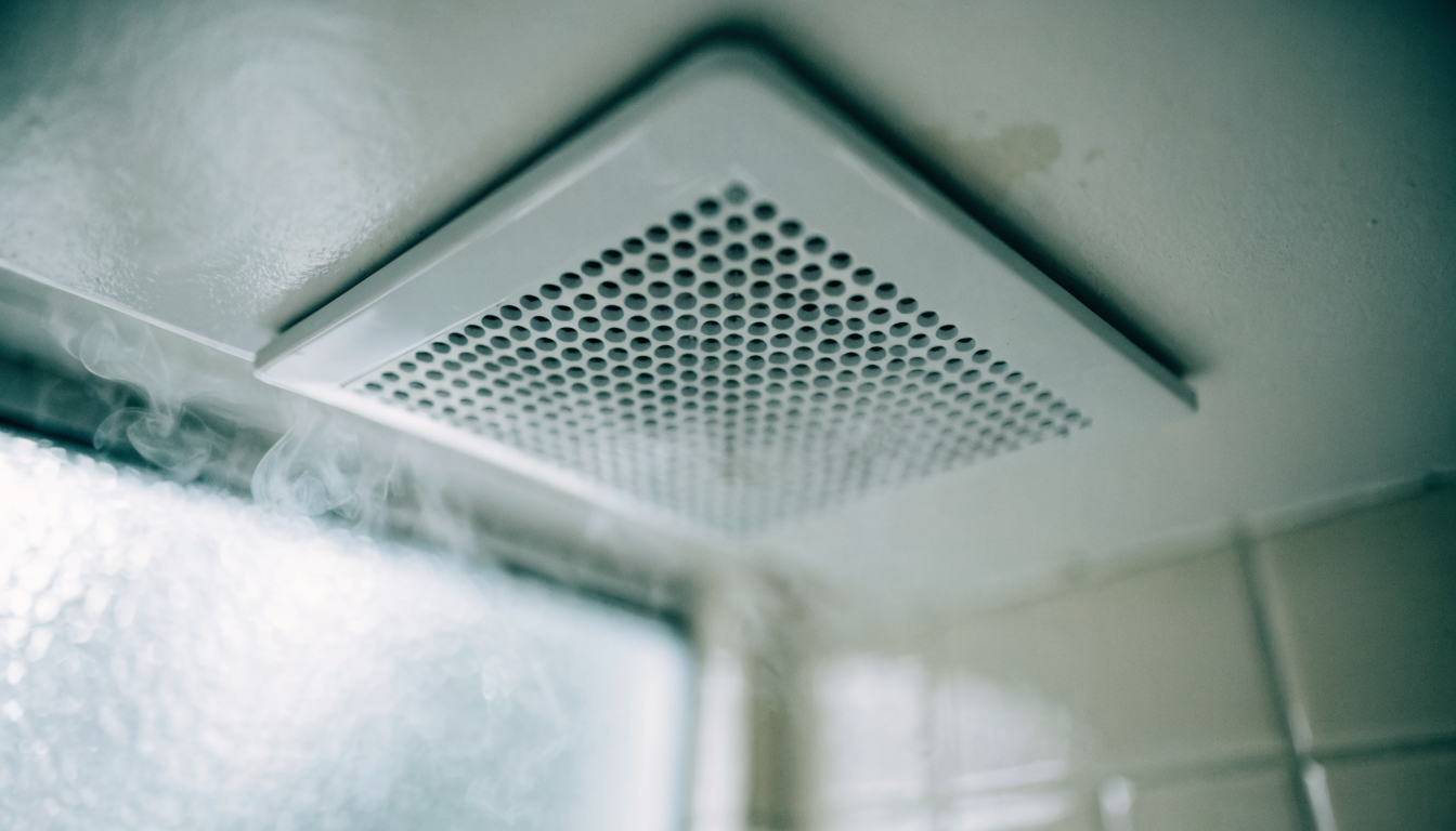 A white square ceiling extractor fan in a tiled bathroom with steam drifting past the grille