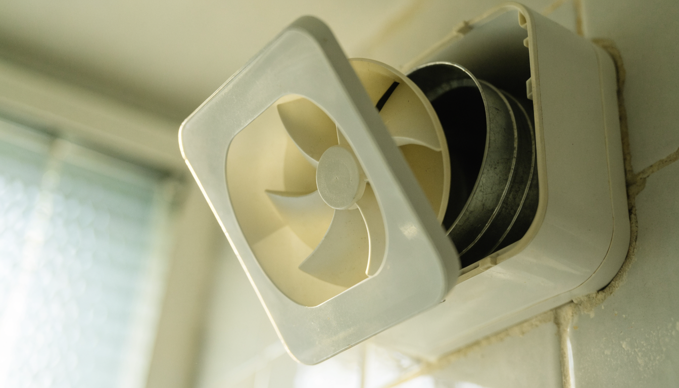 A white bathroom extractor fan on a tiled wall with its front cover unclipped and pulled away, exposing the impeller and the dark duct opening behind it