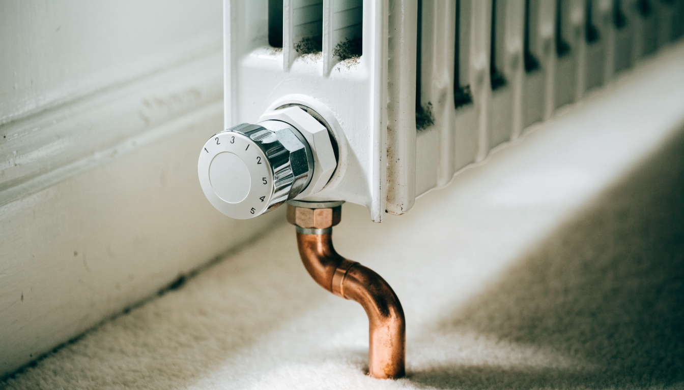 A white thermostatic radiator valve with a numbered dial fitted to the bottom of a white panel radiator, with the copper pipe running down into the floor