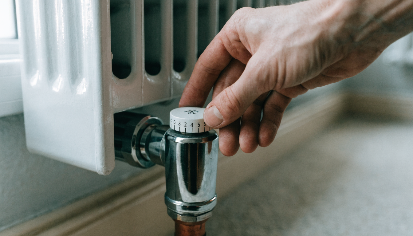 A hand turning the numbered dial of a thermostatic radiator valve down towards the frost setting on a white panel radiator